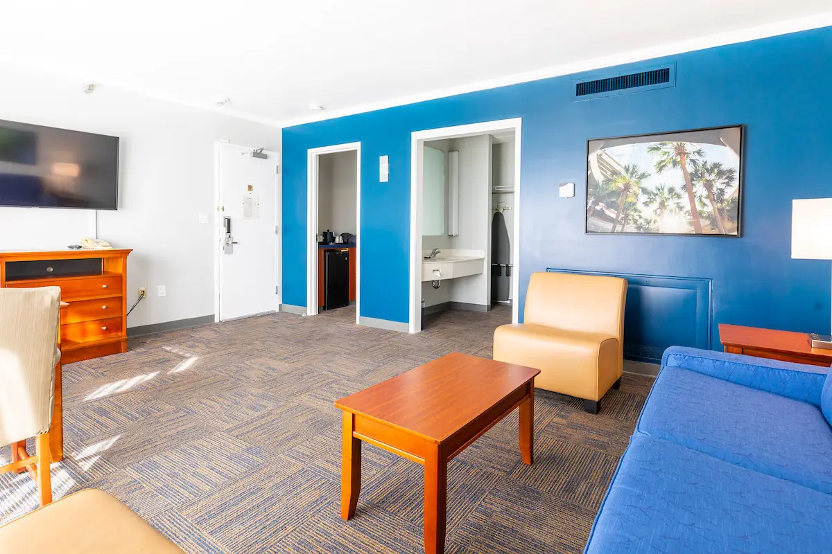 A hotel room with a blue wall, a TV on a dresser, a yellow chair, a blue couch, and a wooden coffee table.