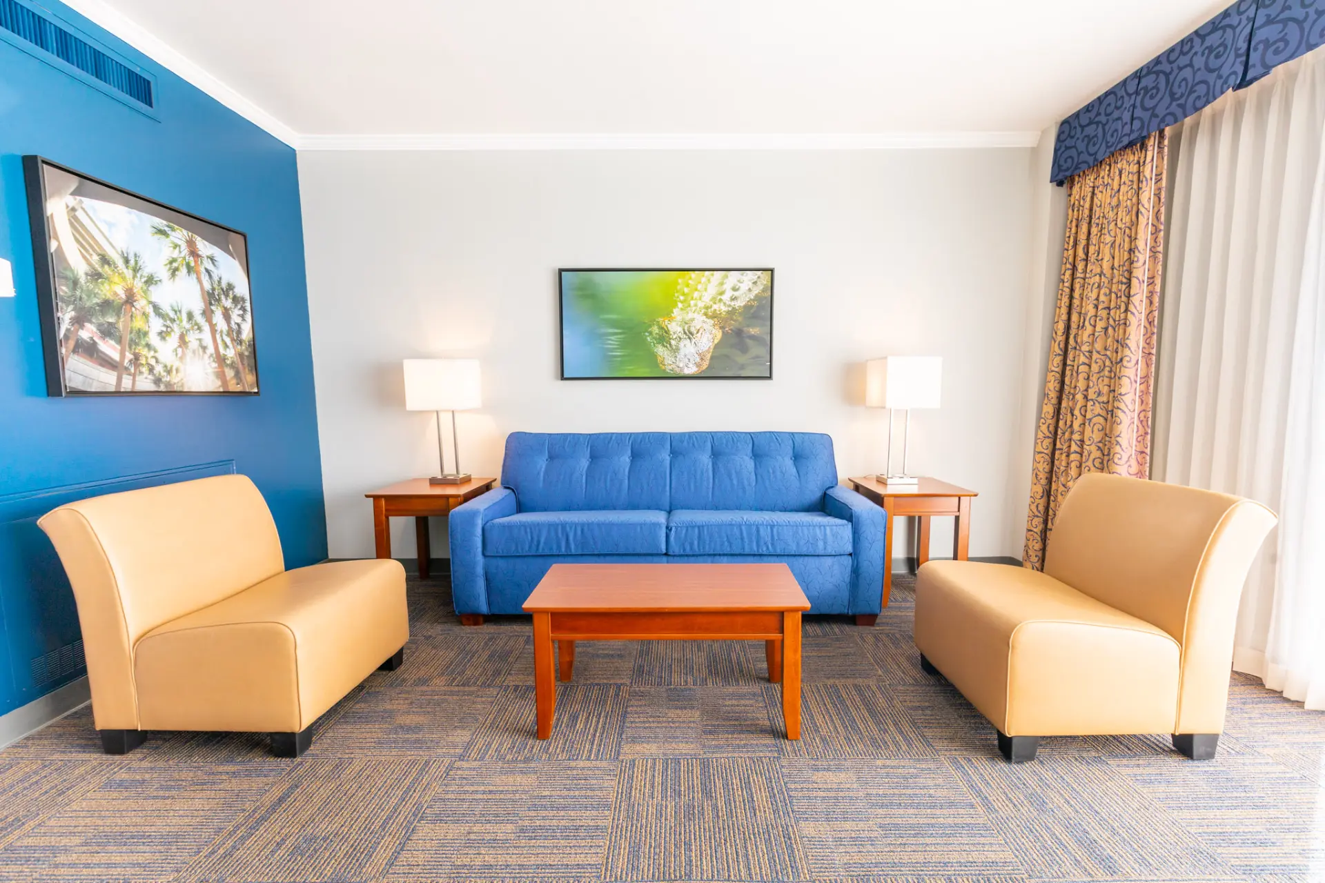 Blue couch, yellow couch, coffee table, wall art, two side tables, lamps, curtains, window, white ceiling, blue wall, carpeted floor.