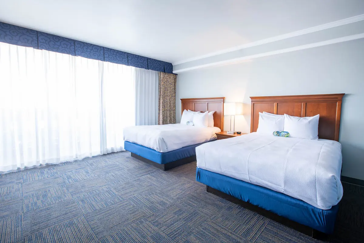 Two twin beds in a hotel room with white sheets, blue bed skirts, and a brown headboard. A window with a blue valance and white curtains is on the left.