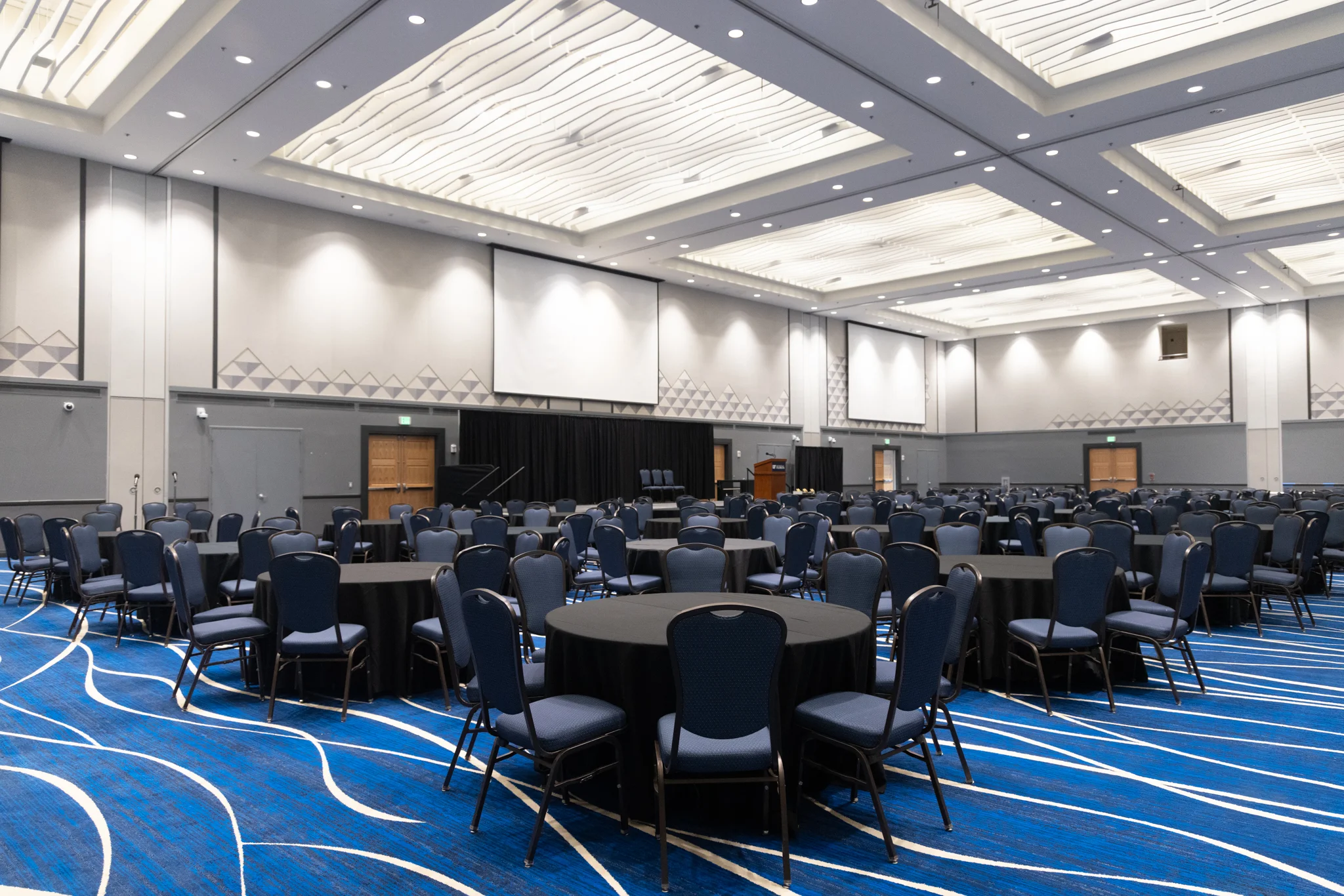 A large conference room with a blue carpet, white ceiling, and rows of tables and chairs.