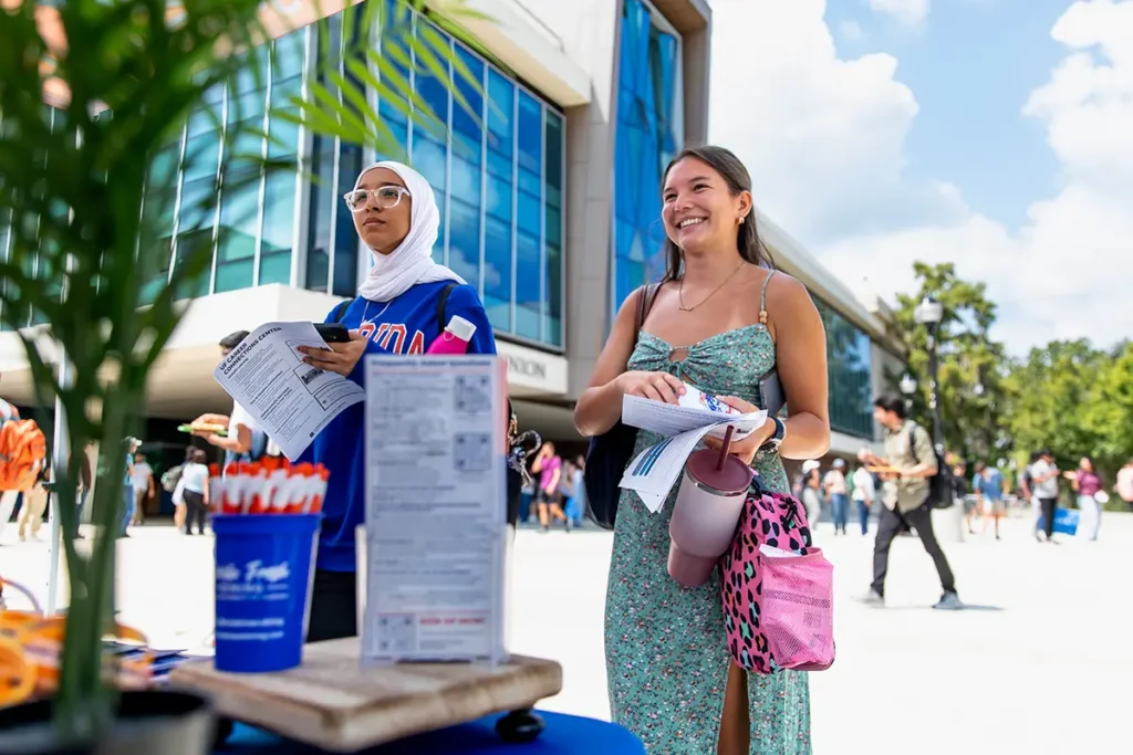 Two women standing outside a building, one wearing a hijab and holding papers, the other in a dress and carrying a pink bag, with a table and sign in the foreground.