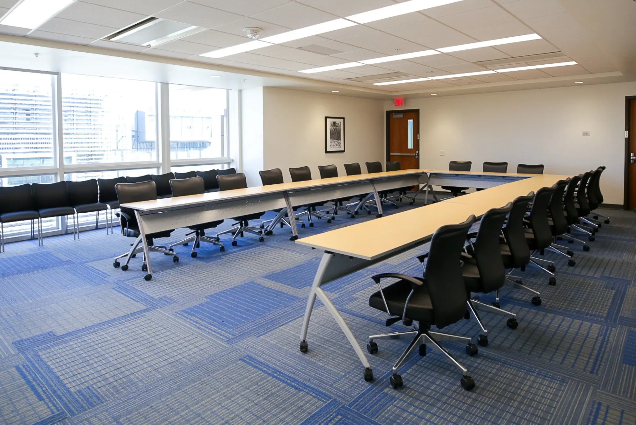 A conference room with a long table and chairs, featuring windows, a door, and a white ceiling with fluorescent lights.