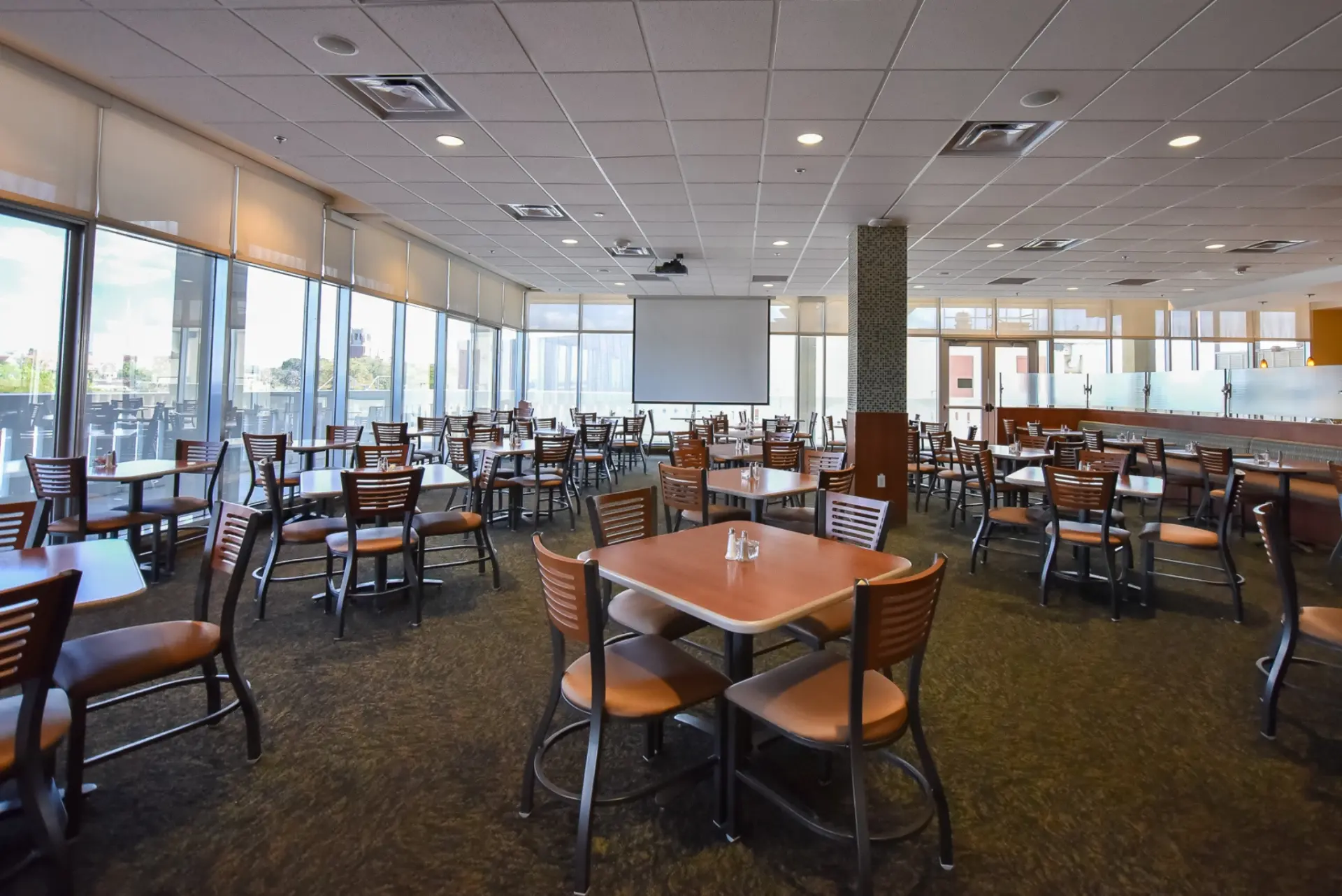 A spacious dining area with a square table and chairs, featuring a projector screen on the wall and large windows offering a view of the outdoors.