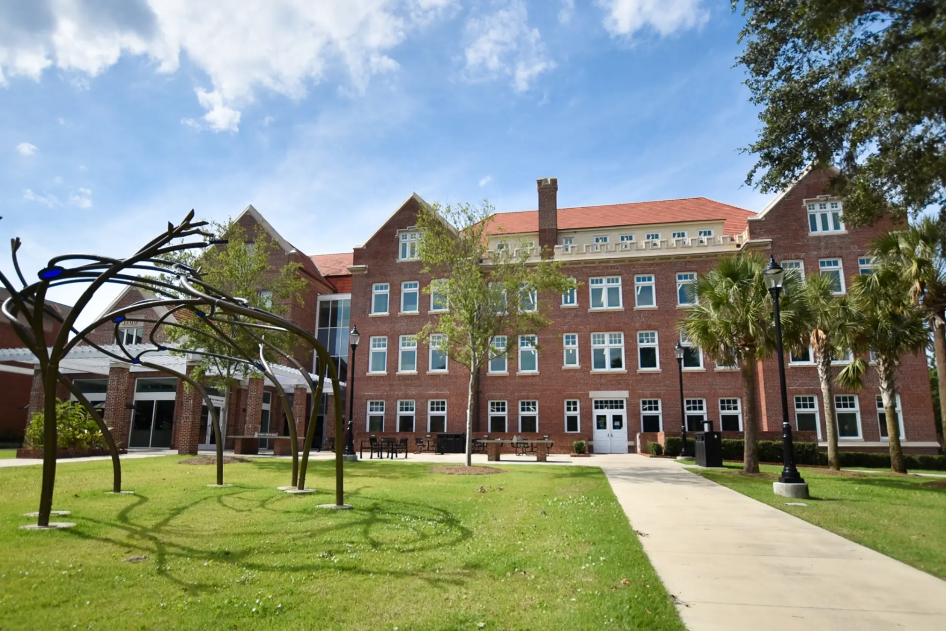 A large red brick building with many windows, a red roof, and a sidewalk in front, surrounded by trees and grass.