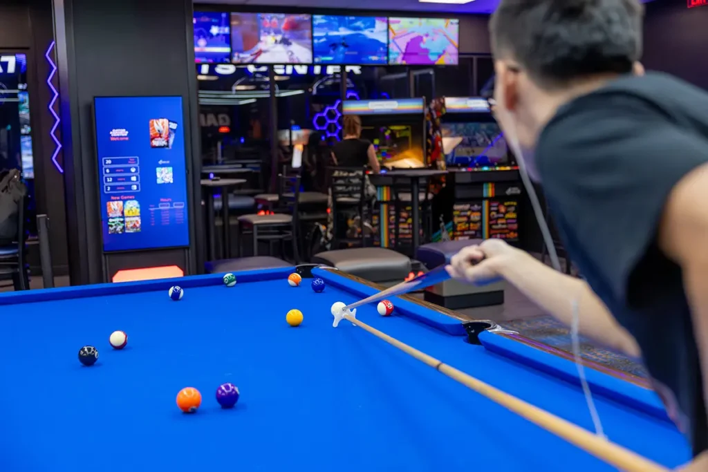 A person playing pool in a game room with a blue table and balls scattered about.