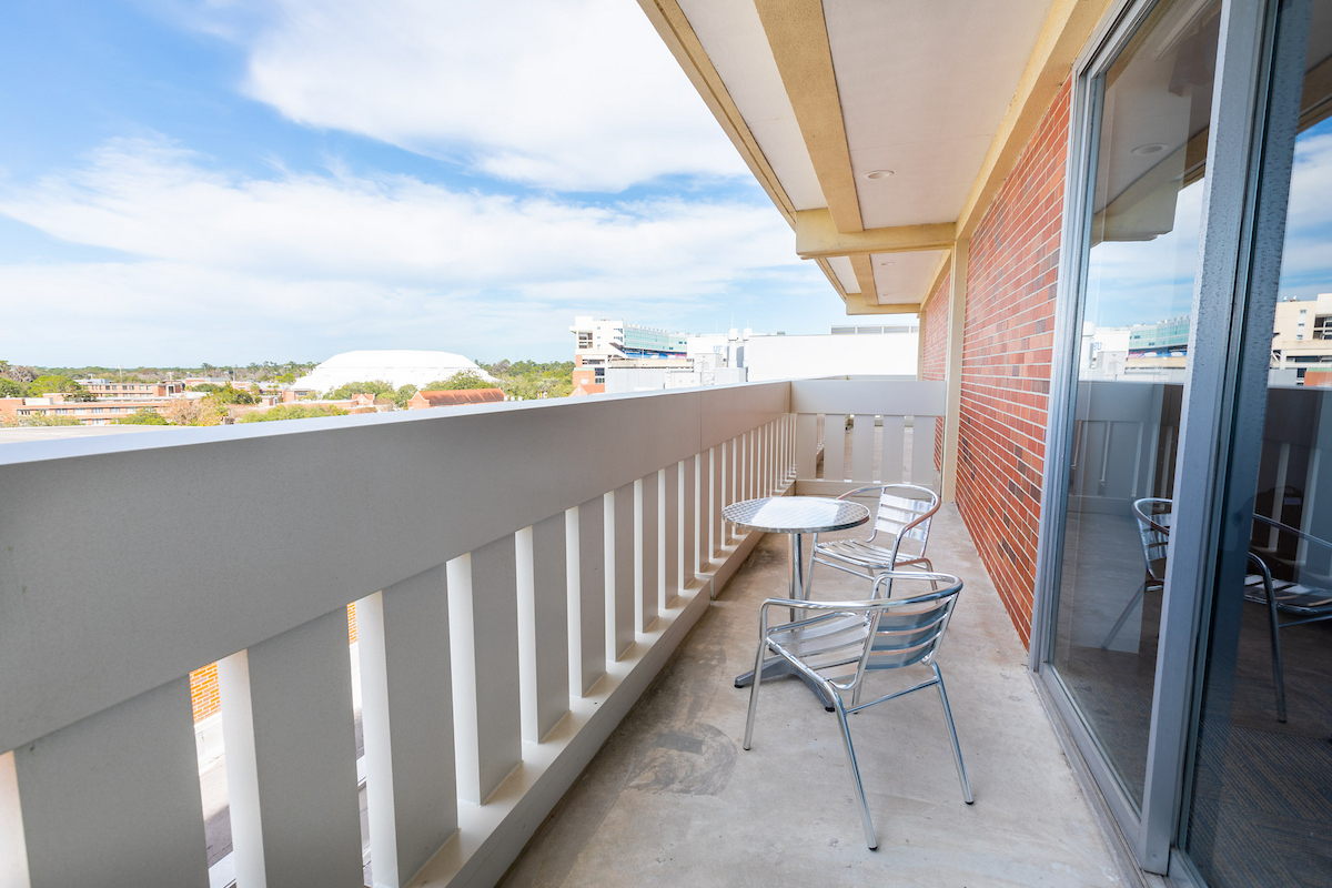 A balcony with a table and chairs, offering a view of a building and a blue sky with clouds.