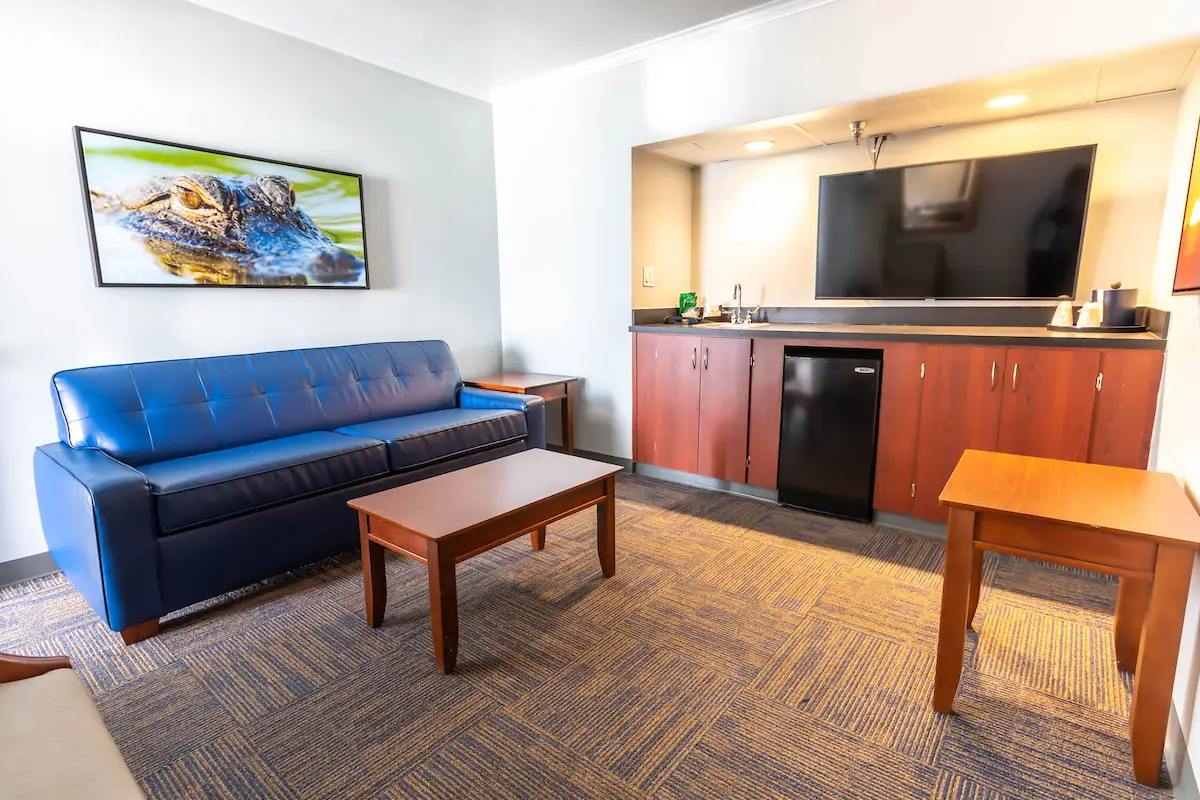 A hotel room with a blue couch, a wooden coffee table, a small refrigerator, a TV, and a painting on the wall.