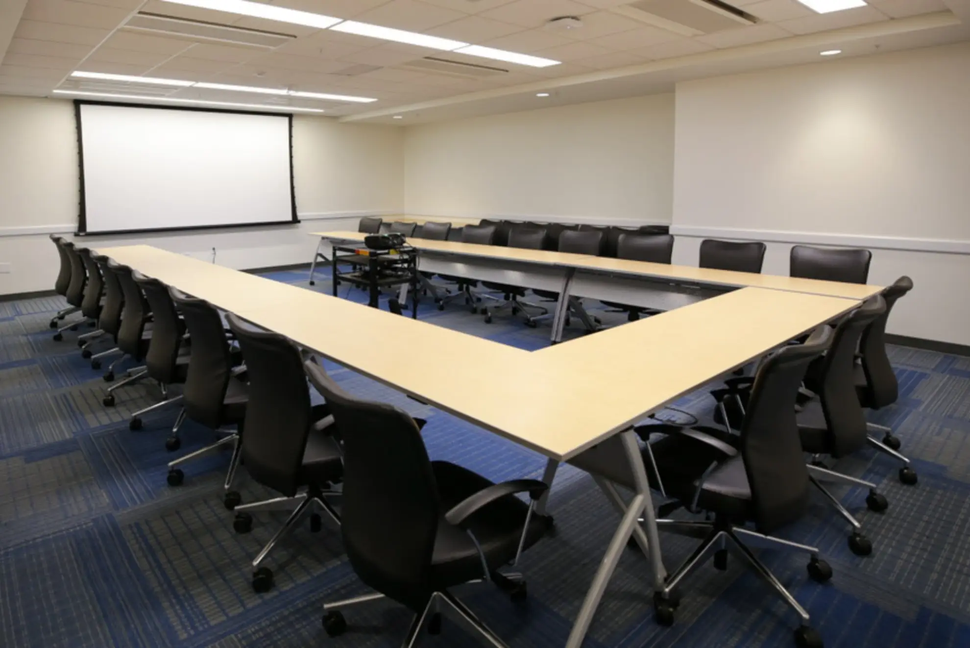 A conference room with a long table and chairs, featuring a projector screen on the wall.