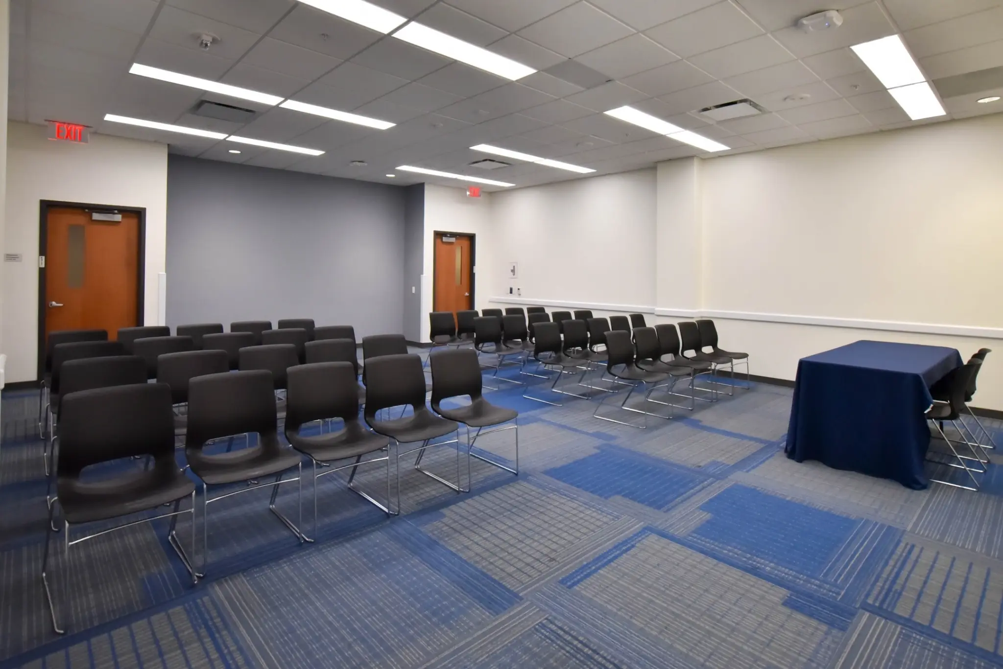 A conference room with a table and chairs, featuring a door, lights, and a blue and gray carpet.