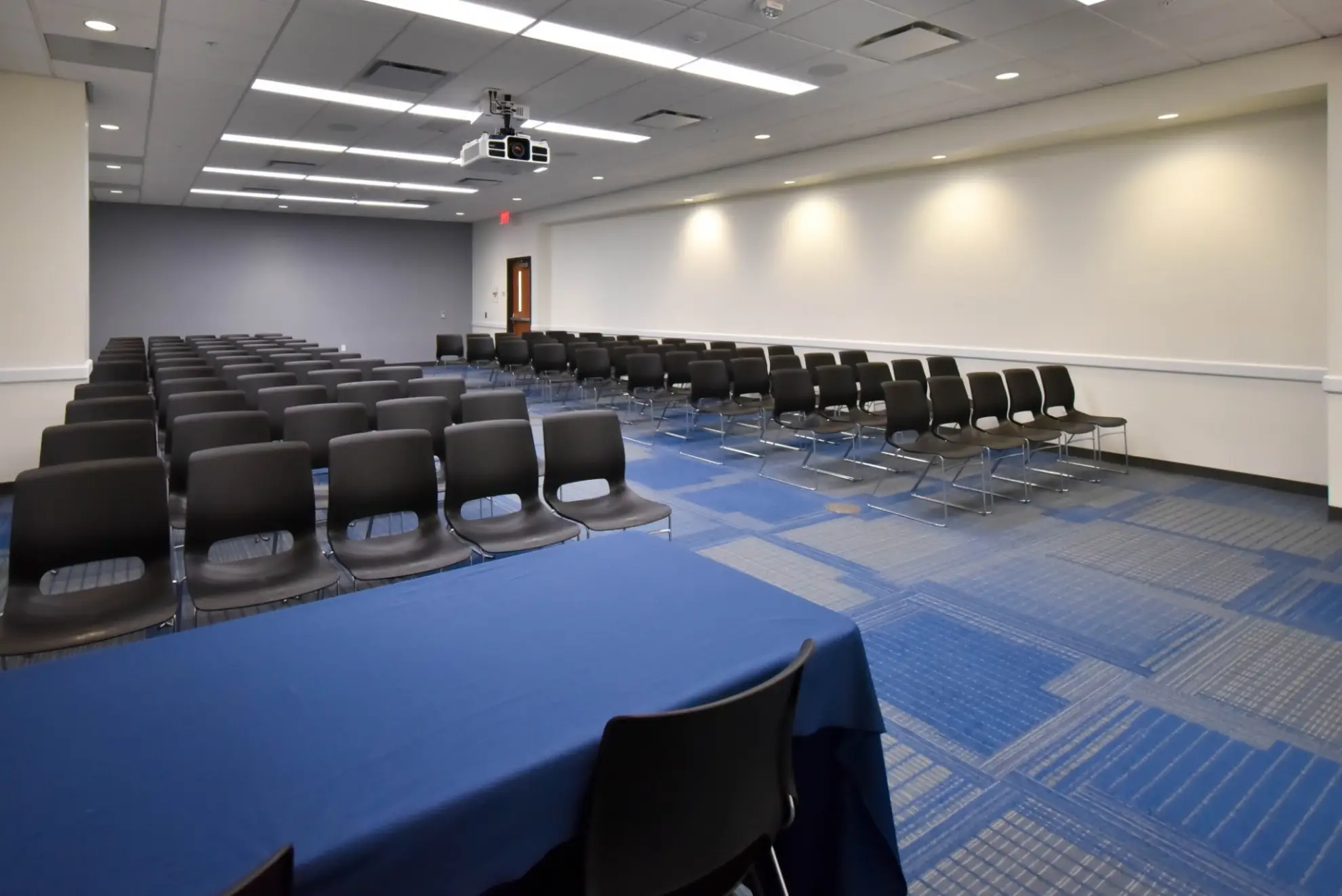 A large conference room with rows of black chairs, a blue table, and a projector screen.