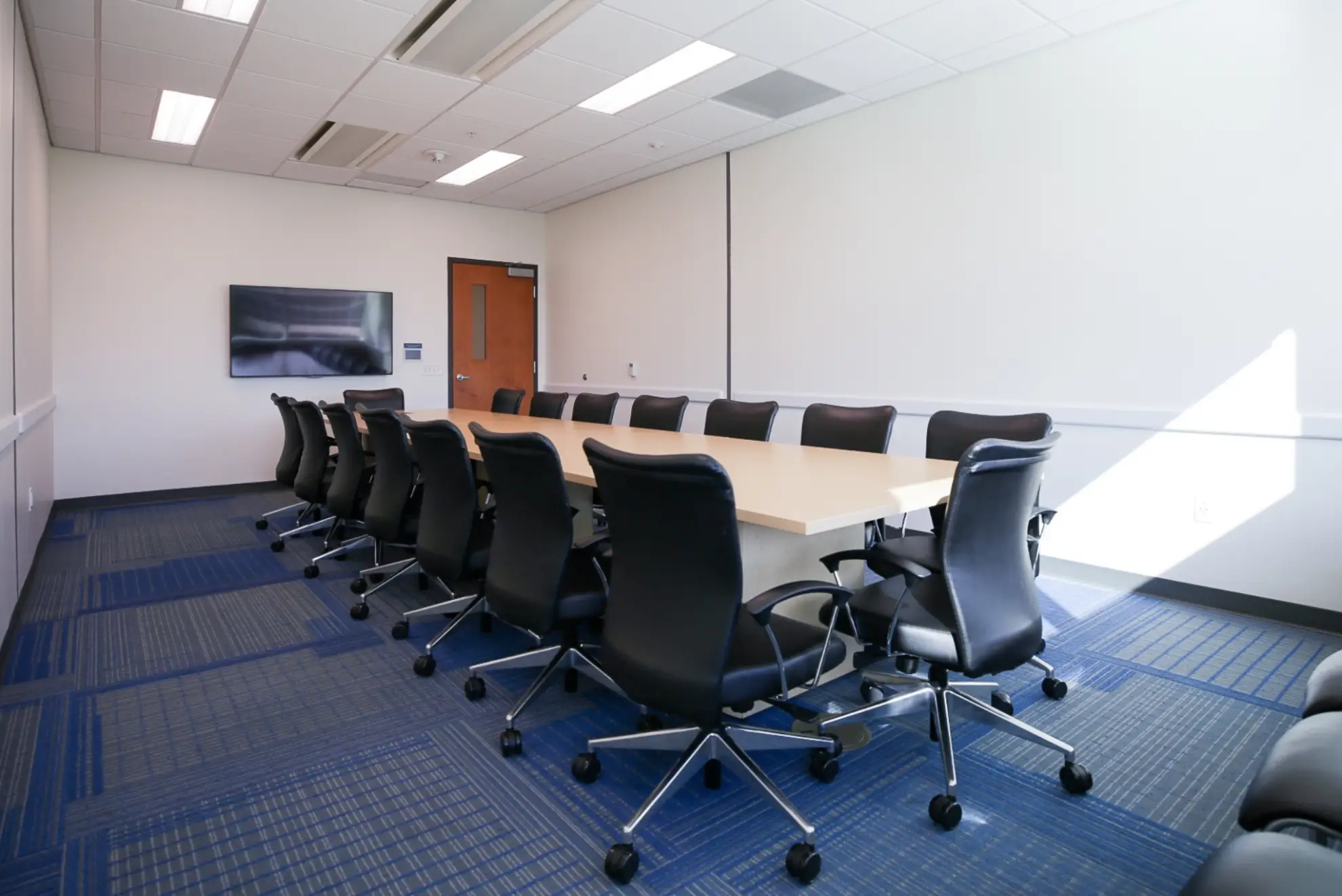 A conference room with a long table and chairs, featuring a TV on the wall and a door.