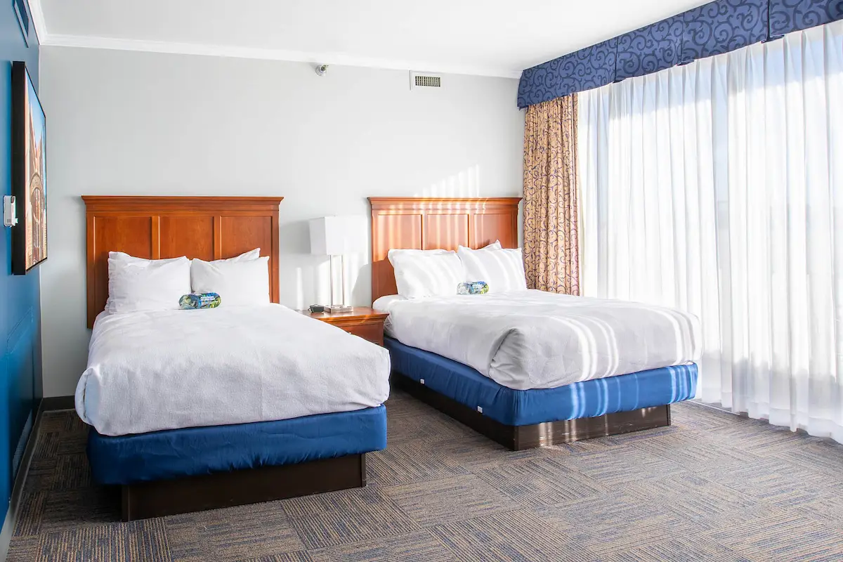 two beds in a hotel room with white sheets and blue blankets, a wooden headboard, and a window with white curtains.